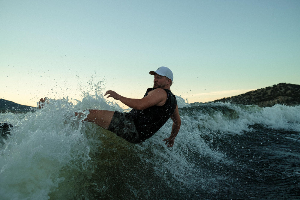A man in a white Suay Apex cap and black tank top wakeboarding, suspended mid-air over a large wave, with a sunset sky and mountains.