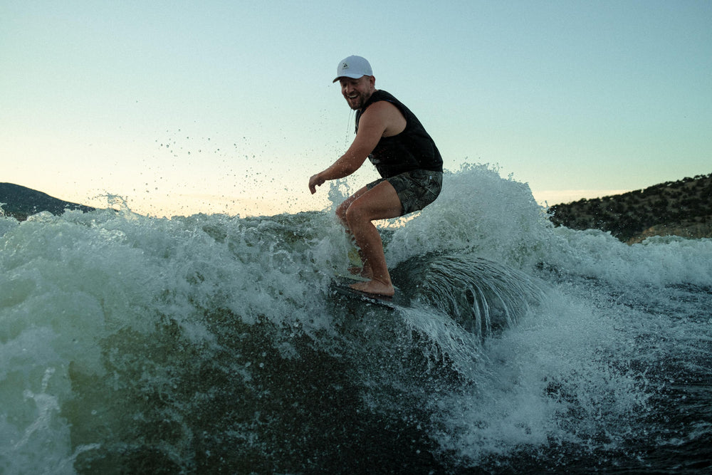 A smiling man wakesurfing on a large wave, wearing a white baseball cap, against a golden-hour sky and blurred hillside.