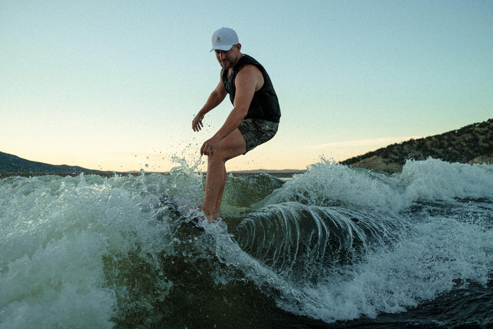 A man in a white Suay cap and black life vest actively surfs a wave, kicking up water against a backdrop of distant mountains and a warm, gradient sky.