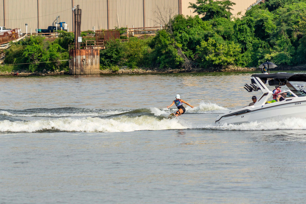 A woman wearing a colorful top and white cap rides a wakesurf board, creating a wake behind a white boat on a sunny river.