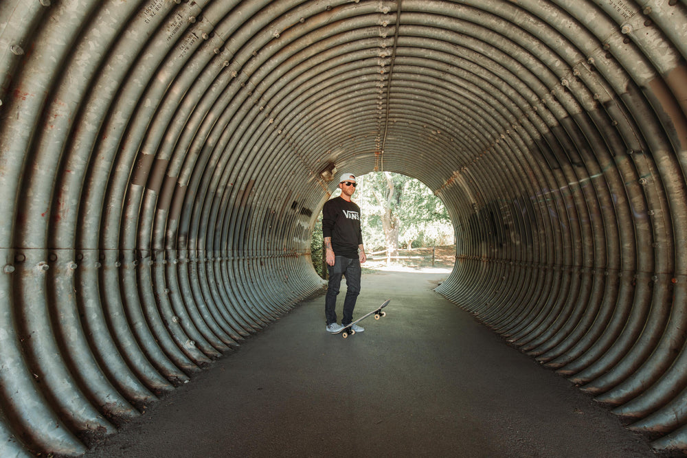 A man wearing a white Suay cap and black long-sleeve shirt stands with a skateboard in a corrugated metal tunnel, looking cool and relaxed.