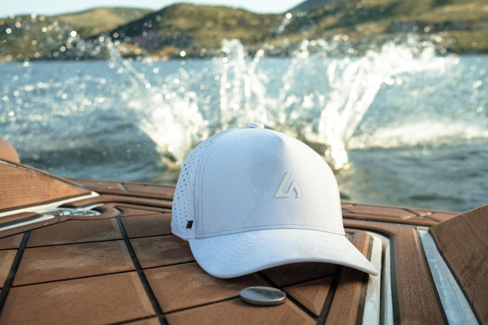 A white Suay Apex hat rests on a boat's teak deck, framed by dynamic water splashes and distant mountains, showcasing an active outdoor lifestyle.