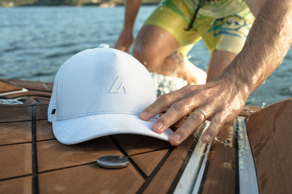 A person's wet hand reaches for a white Suay Apex hat resting on a textured wooden boat deck, with blurred water and legs visible in the background.