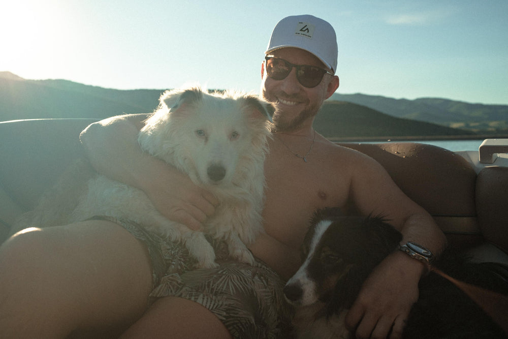 A smiling man in a white Suay hat and sunglasses, embracing two dogs on a boat, against a backdrop of mountains and water during golden hour.