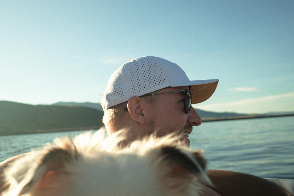 A man in a white perforated hat and sunglasses smiles, looking over a lake at sunset, with a blurred dog partially visible in the foreground.