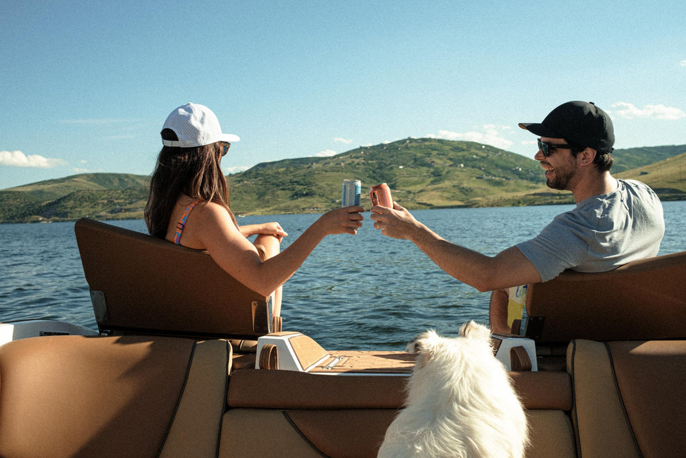 Two people wearing Suay hats are seated on a boat on a sunny lake, clinking beverage cans, with a white dog nearby and green hills in the background.
