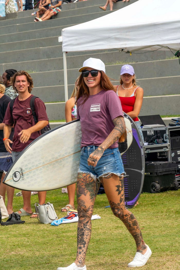 A smiling woman with tattoos and sunglasses wears a white cap while walking on grass, holding a surfboard at an outdoor event.