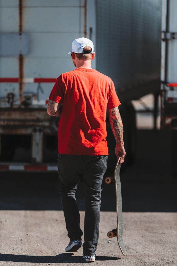 A man wearing a white Suay cap and red t-shirt walks away, holding a skateboard, against a sunlit, blurred industrial trailer background.