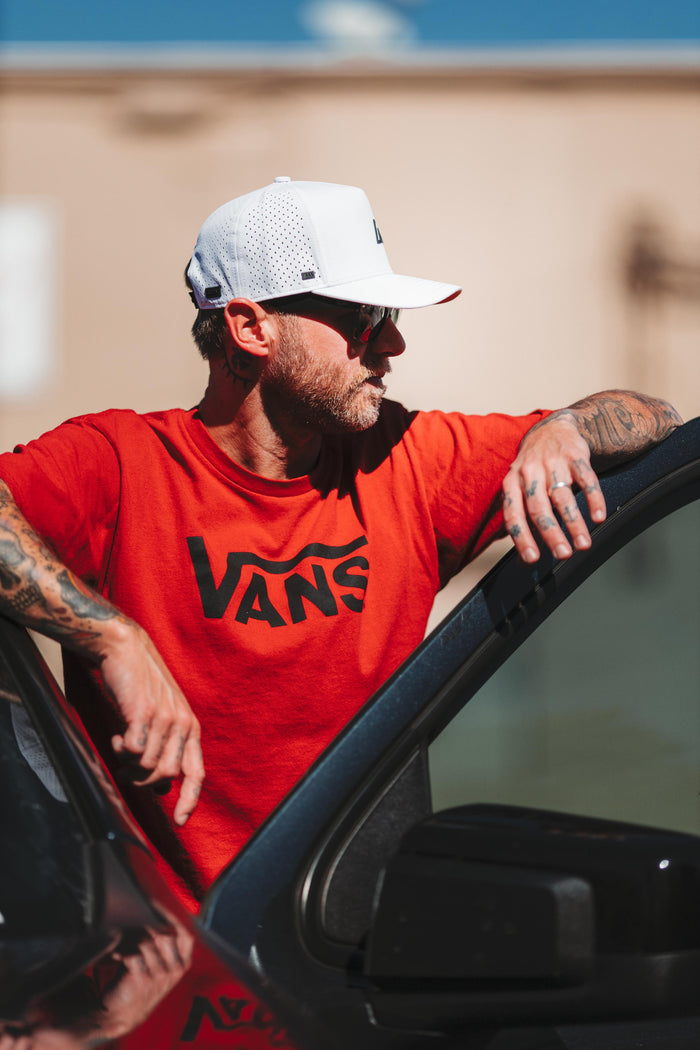 A tattooed man with a beard and sunglasses wears a white Suay Apex hat and a red Vans t-shirt, leaning against a dark vehicle under bright natural light.