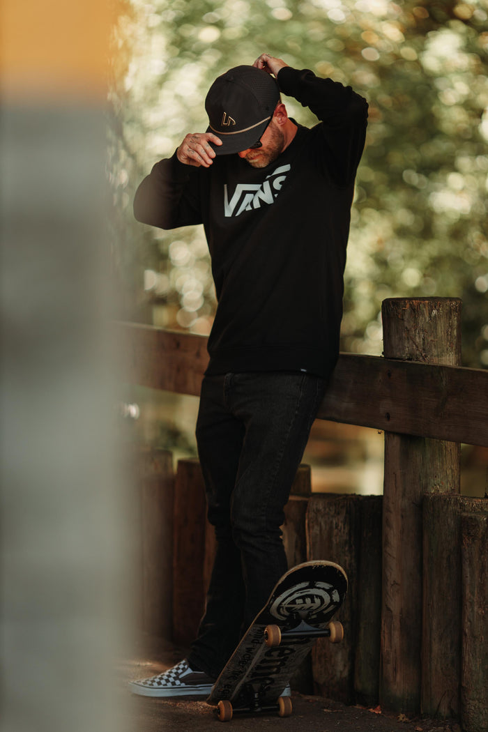 A man in a black Suay Drift hat and black clothing leans casually on a wooden fence next to a skateboard in a warm, outdoor setting.