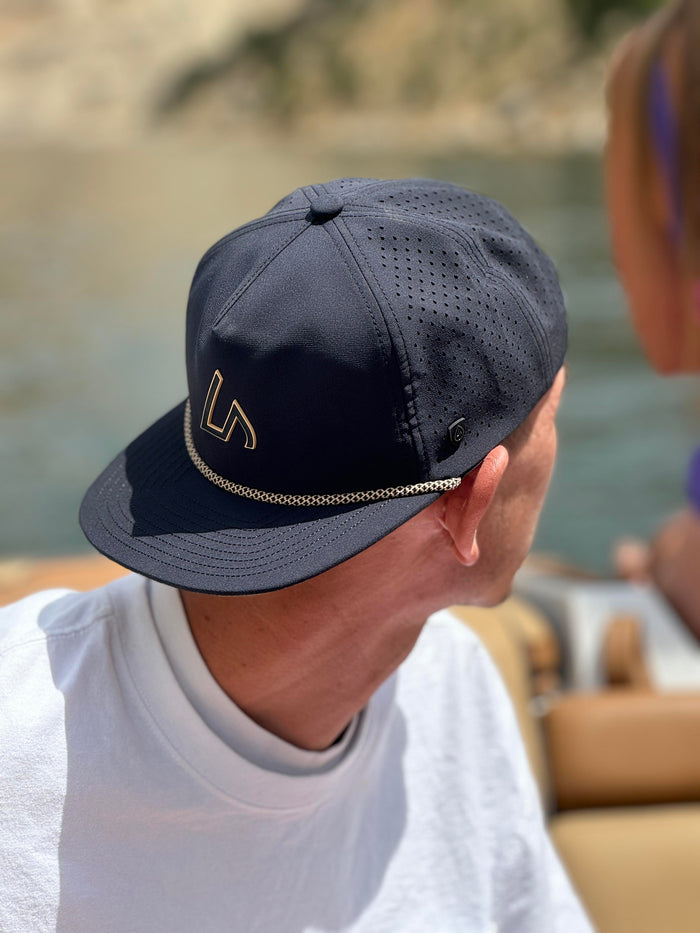 A person wearing a black Suay Drift hat with a gold logo and rope detail looks over blurred water and distant hills.