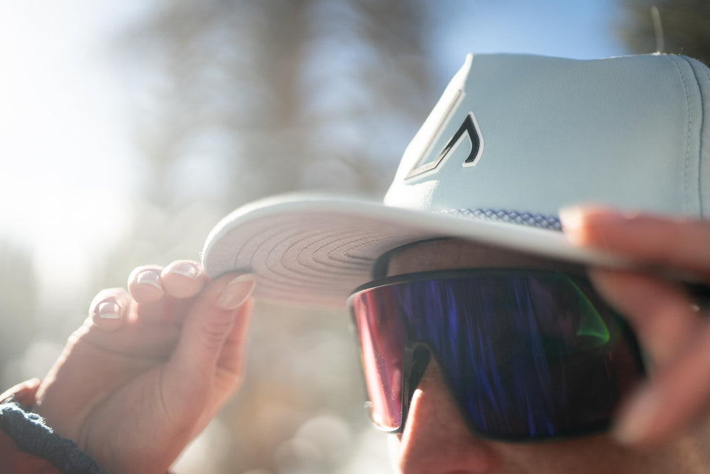 A person wearing a mint Suay Drift hat and mirrored sunglasses is adjusting the hat's brim outdoors, bathed in bright sunlight.