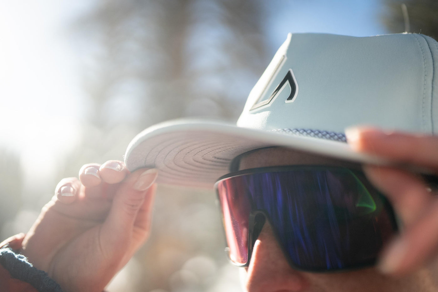 A person wearing a mint Suay Drift hat and mirrored sunglasses is adjusting the hat's brim outdoors, bathed in bright sunlight.