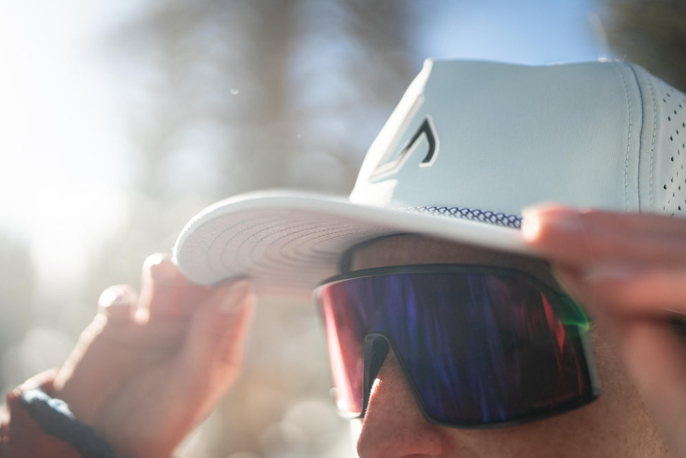 A person adjusts a light blue drift-style hat with a rope detail, while wearing reflective sunglasses in bright outdoor sunlight.