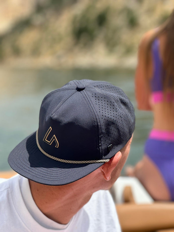 A person wears a navy performance hat with a gold Suay logo and rope detail while relaxing on a boat, with a blurred lake in the background.