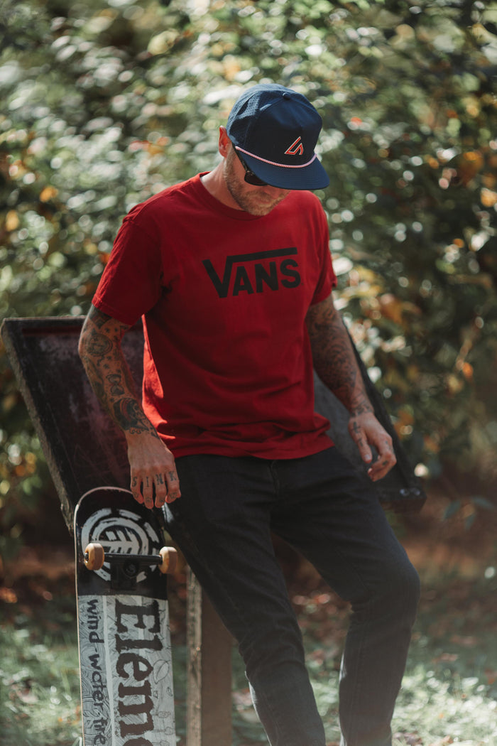 A person wearing a navy Suay Drift hat with a red rope detail and a red Vans t-shirt, stands outdoors with a skateboard, surrounded by blurred greenery.