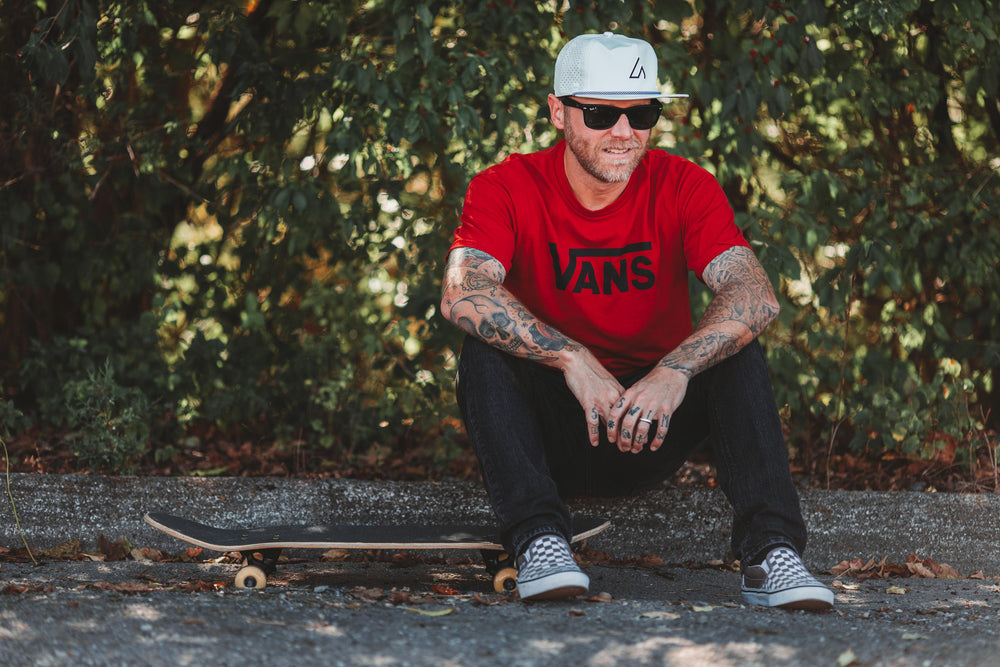 A bearded man in sunglasses, a white Suay hat, and a red Vans t-shirt sits casually on the ground next to a skateboard, looking right and smiling slightly, against a blurred green leafy background.