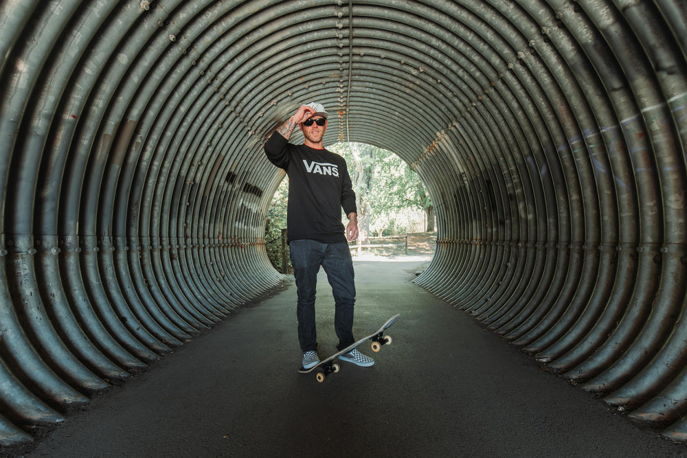 A male skateboarder wearing a white Suay hat and Vans long-sleeve shirt stands in a metal tunnel, looking towards the camera with a skateboard at his feet.
