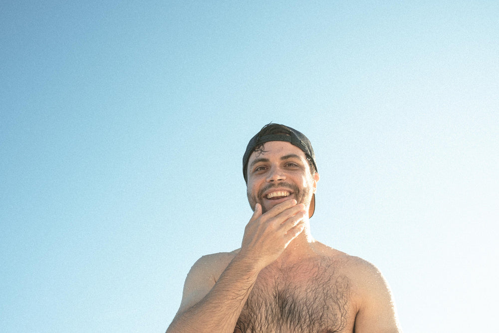 A smiling man wearing a black Suay cap looks up at the camera under a bright blue sky, with natural sunlight highlighting his face and chest.