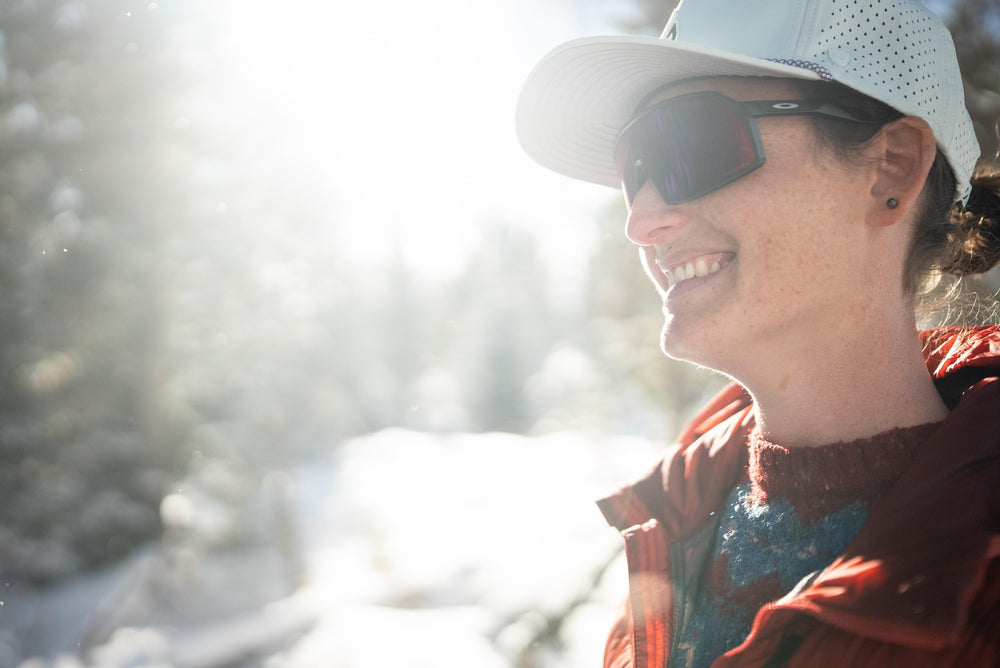 A smiling woman wearing a white Suay performance hat and dark sunglasses is bathed in bright sunlight against a soft, snowy outdoor background.