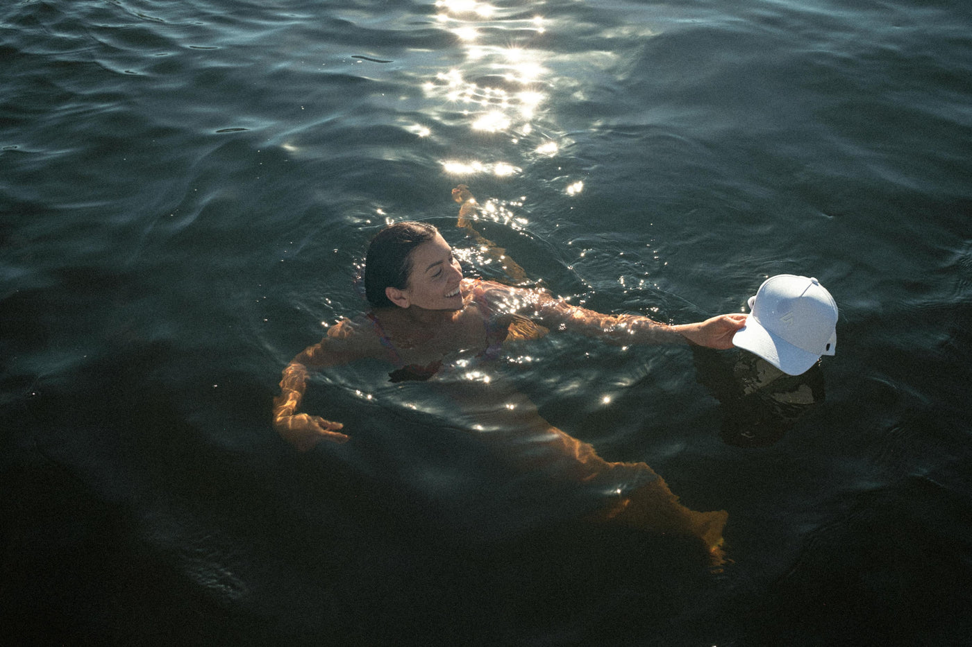 A smiling woman with wet hair swims in dark water, holding a white baseball cap, illuminated by bright sun reflections.