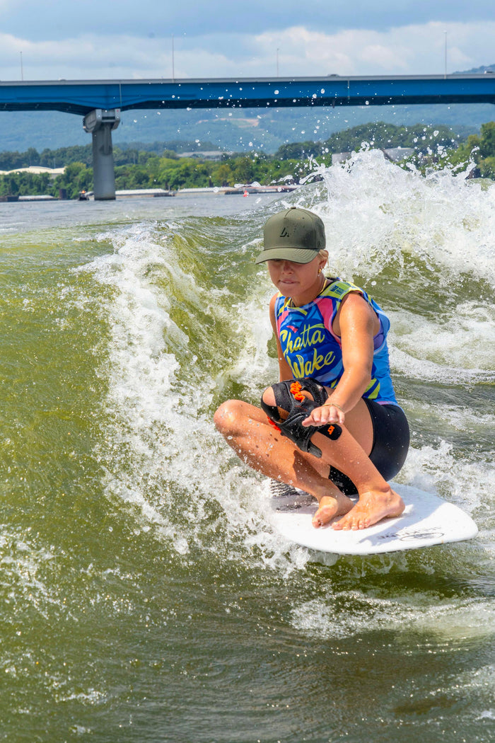 A woman wearing a moss green cap and colorful wetsuit kneels on a wakeboard, riding a foamy green wave with a bridge and hills in the bright background.