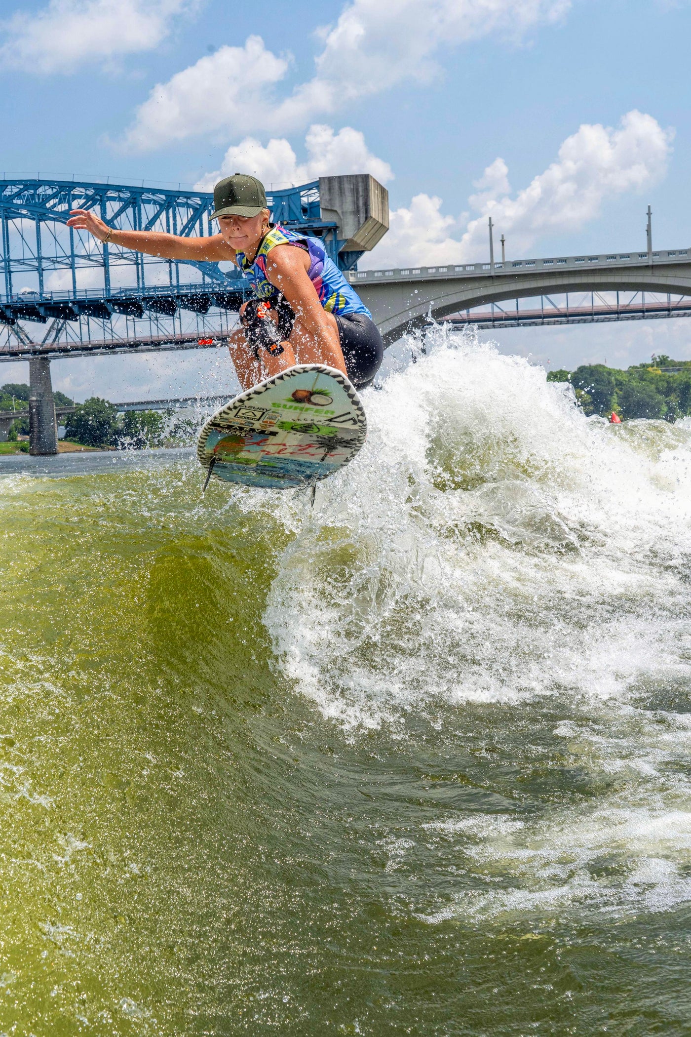 A young person wearing a moss green hat, wakesurfing on a vibrant green wave, with multiple bridges and a bright blue sky in the background.