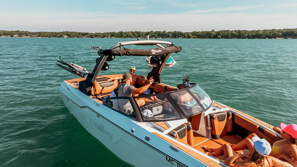 A group of friends enjoying a sunny day on a lake, cruising in a Malibu boat, with several individuals wearing baseball caps.