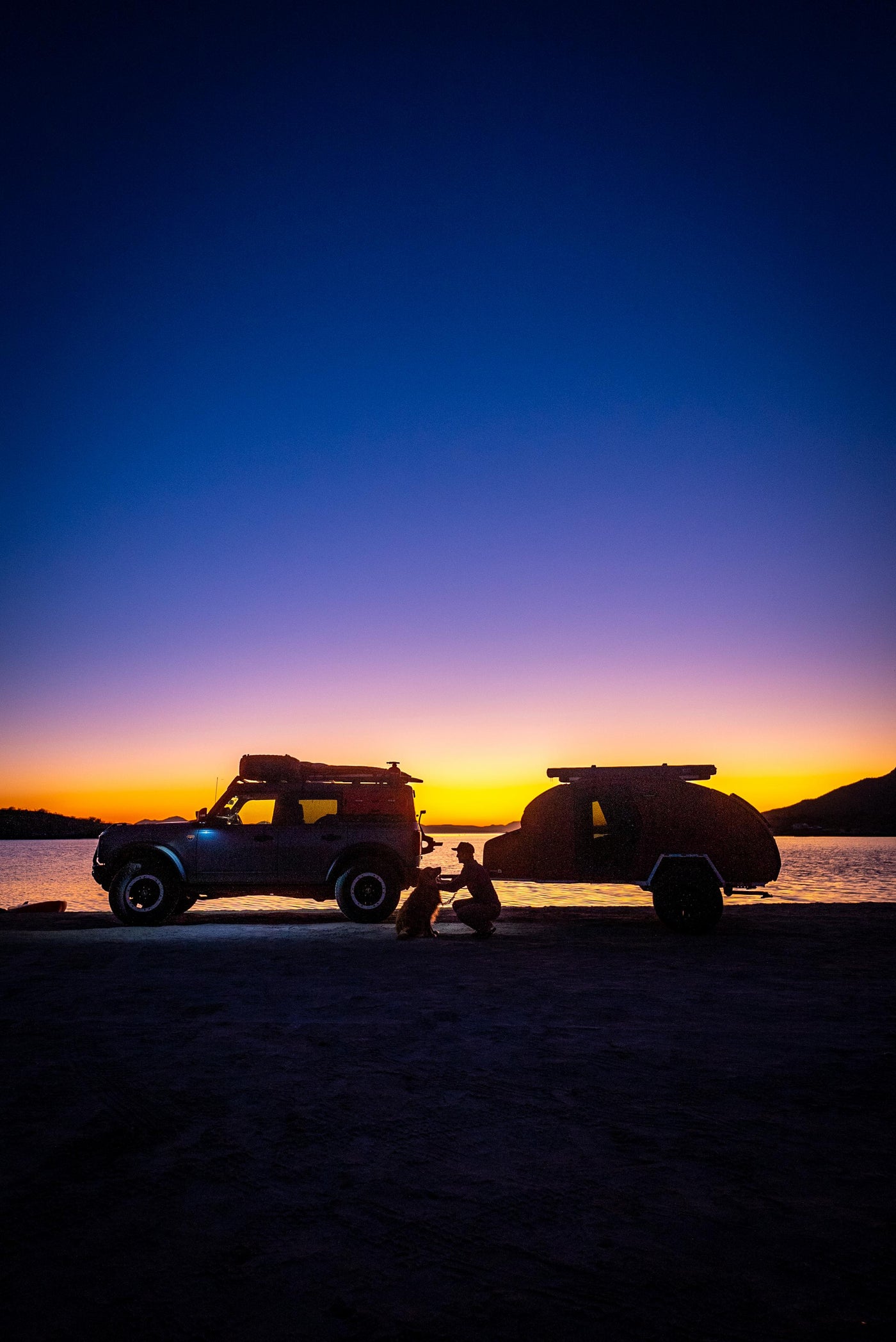 A person kneels with their dog beside an SUV and teardrop trailer by a lake at sunset, silhouetted against a vibrant gradient sky.