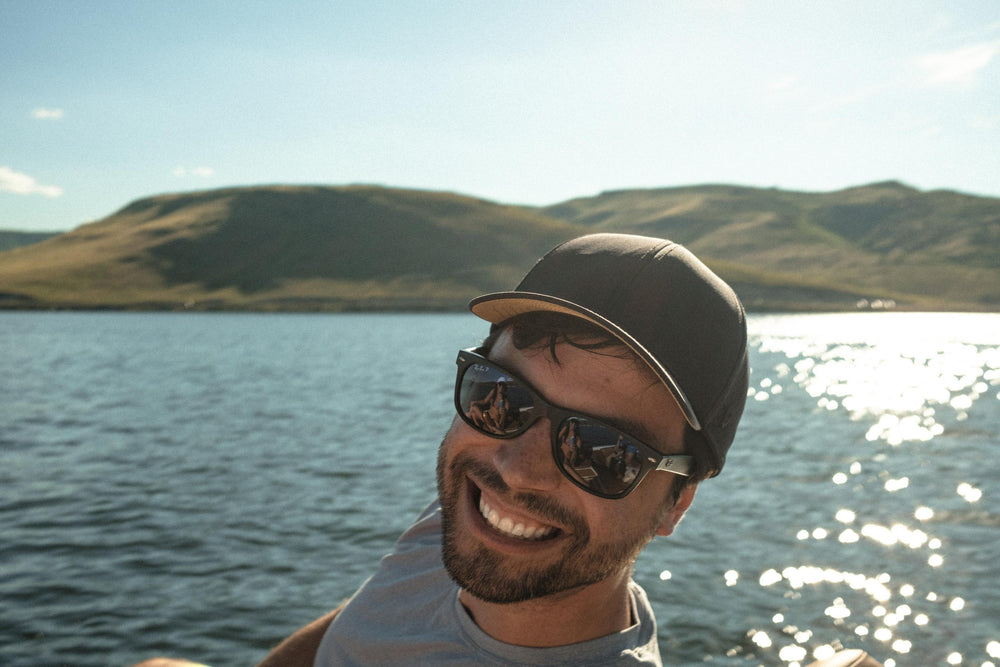 A smiling man wearing a dark cap and sunglasses on a sunny lake, with shimmering water and hills in the background, exuding a relaxed outdoor vibe.