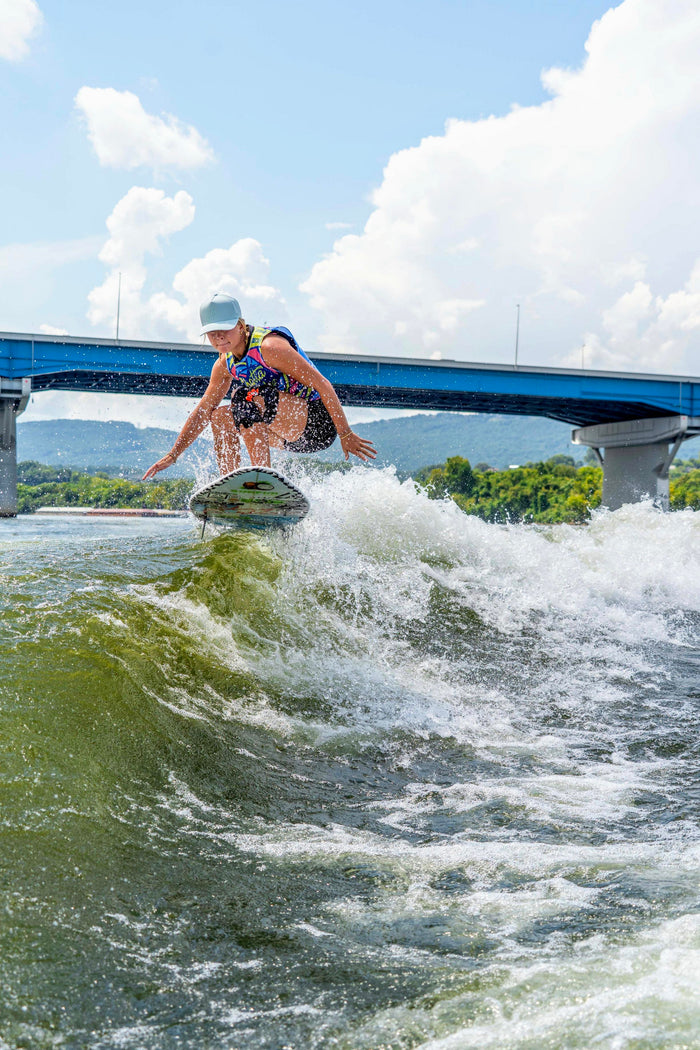A person wearing a light blue Suay cap is wake surfing, creating a large splash on green water under a blue bridge and a bright sky.